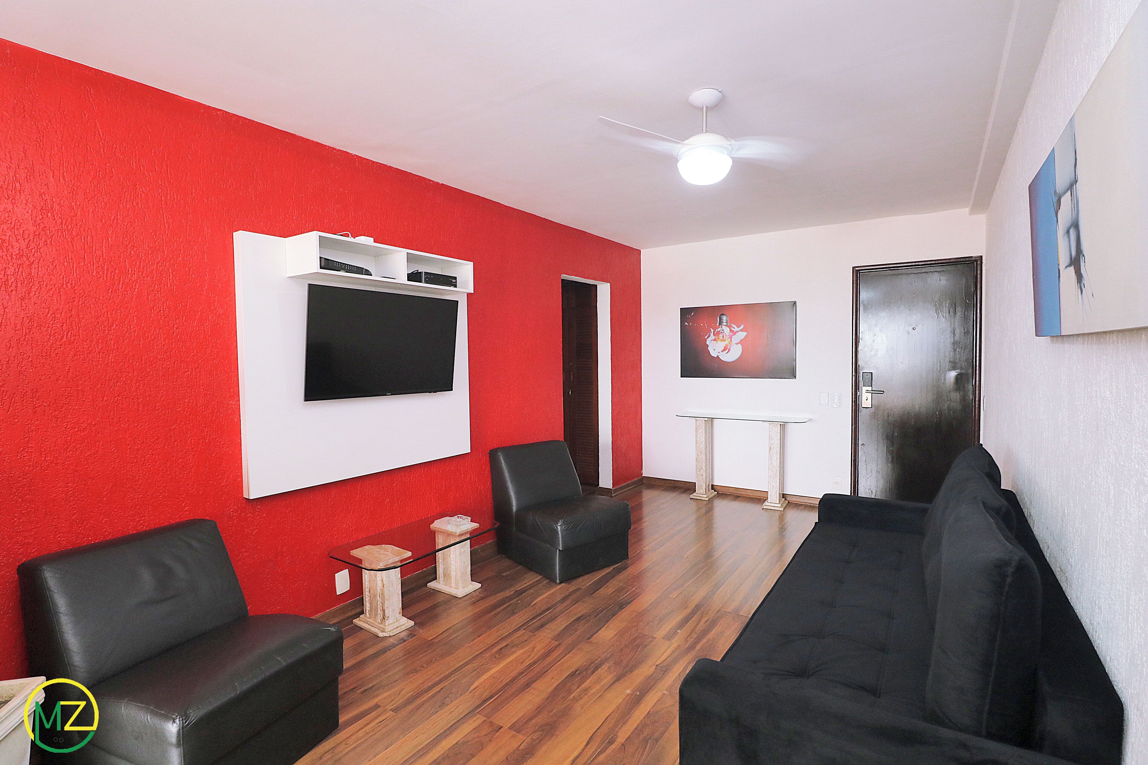 A room with a red wall, a ceiling fan, a flat-screen TV mounted on the wall, a black leather sofa, a black armchair, and a wooden coffee table.