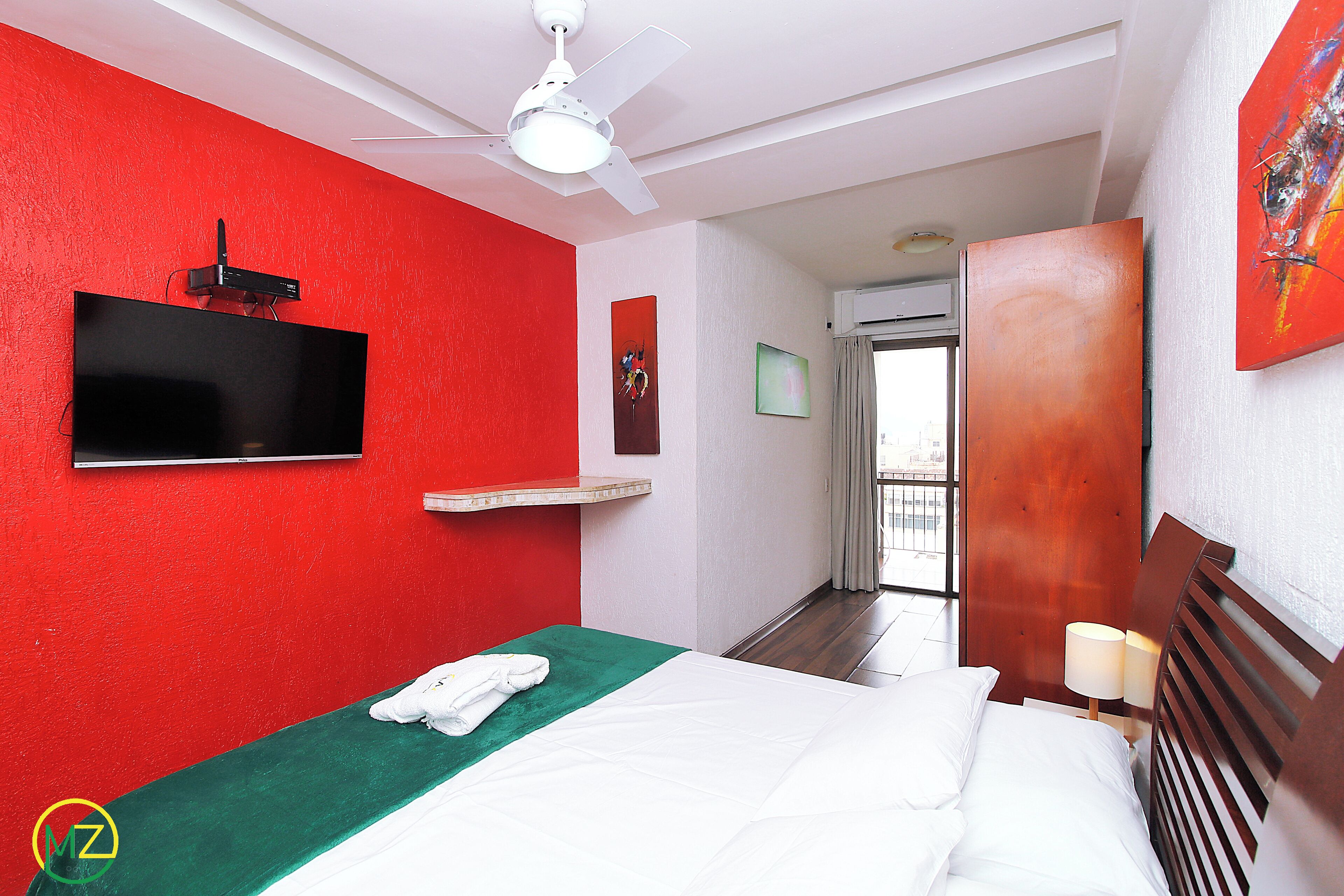 Habitación de hotel con pared de acento roja, un televisor de pantalla plana montado en la pared, cama con ropa blanca, una manta verde y cabecera de madera.