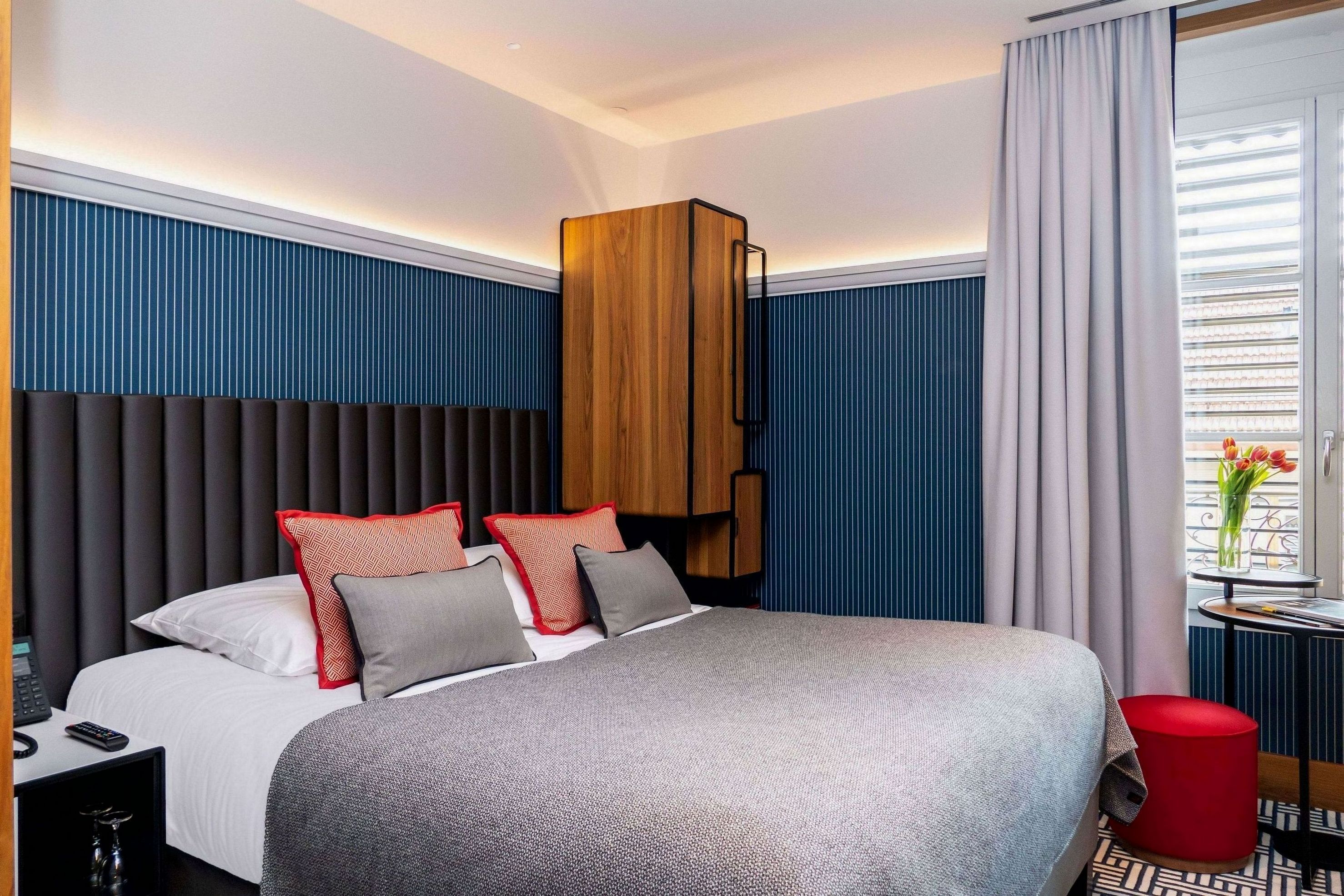 A modern hotel room with a large bed, a wooden headboard, a small table with a vase, and a red stool.
