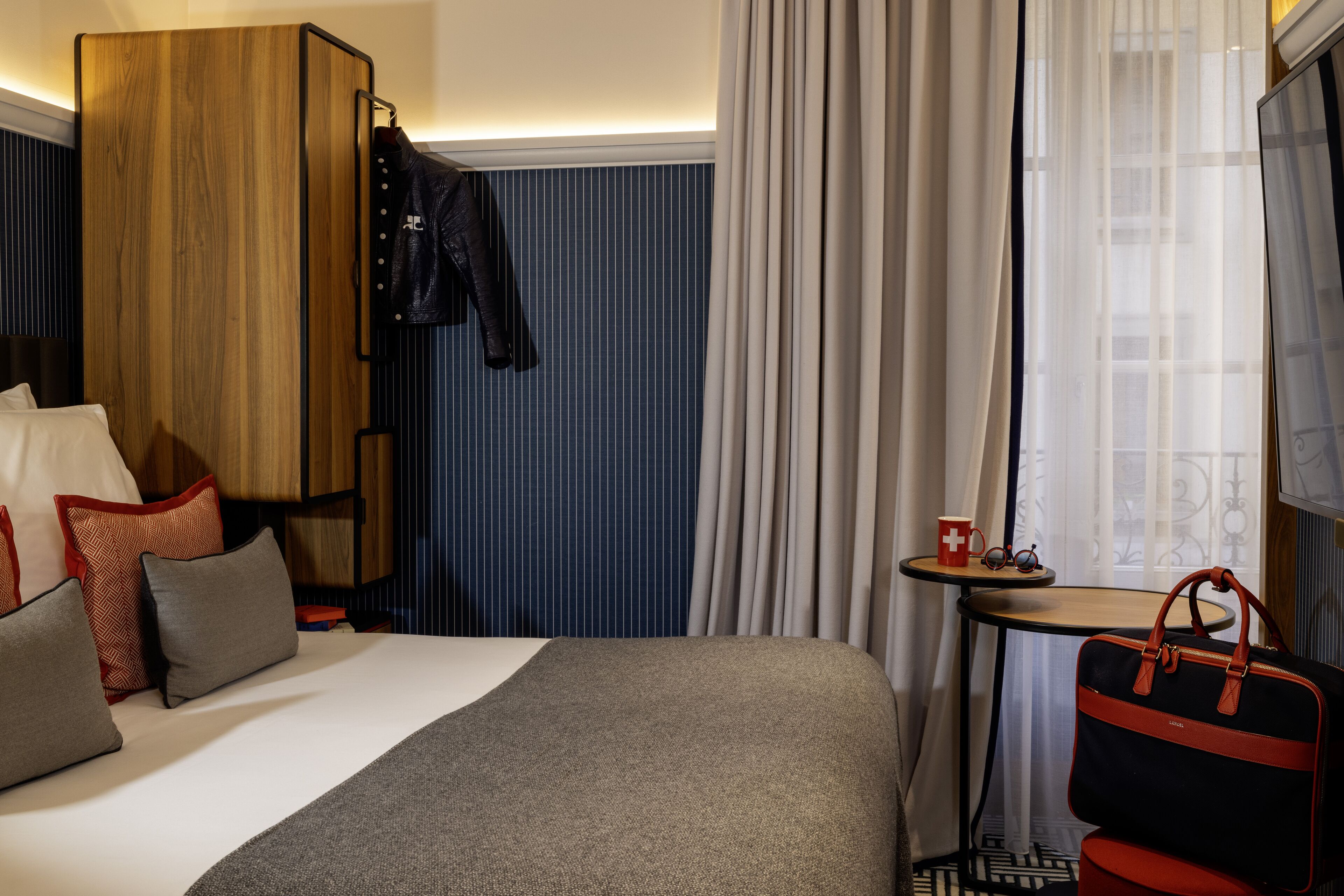 A hotel room with a bed, a wooden wardrobe, a small table with a red mug, and a suitcase.