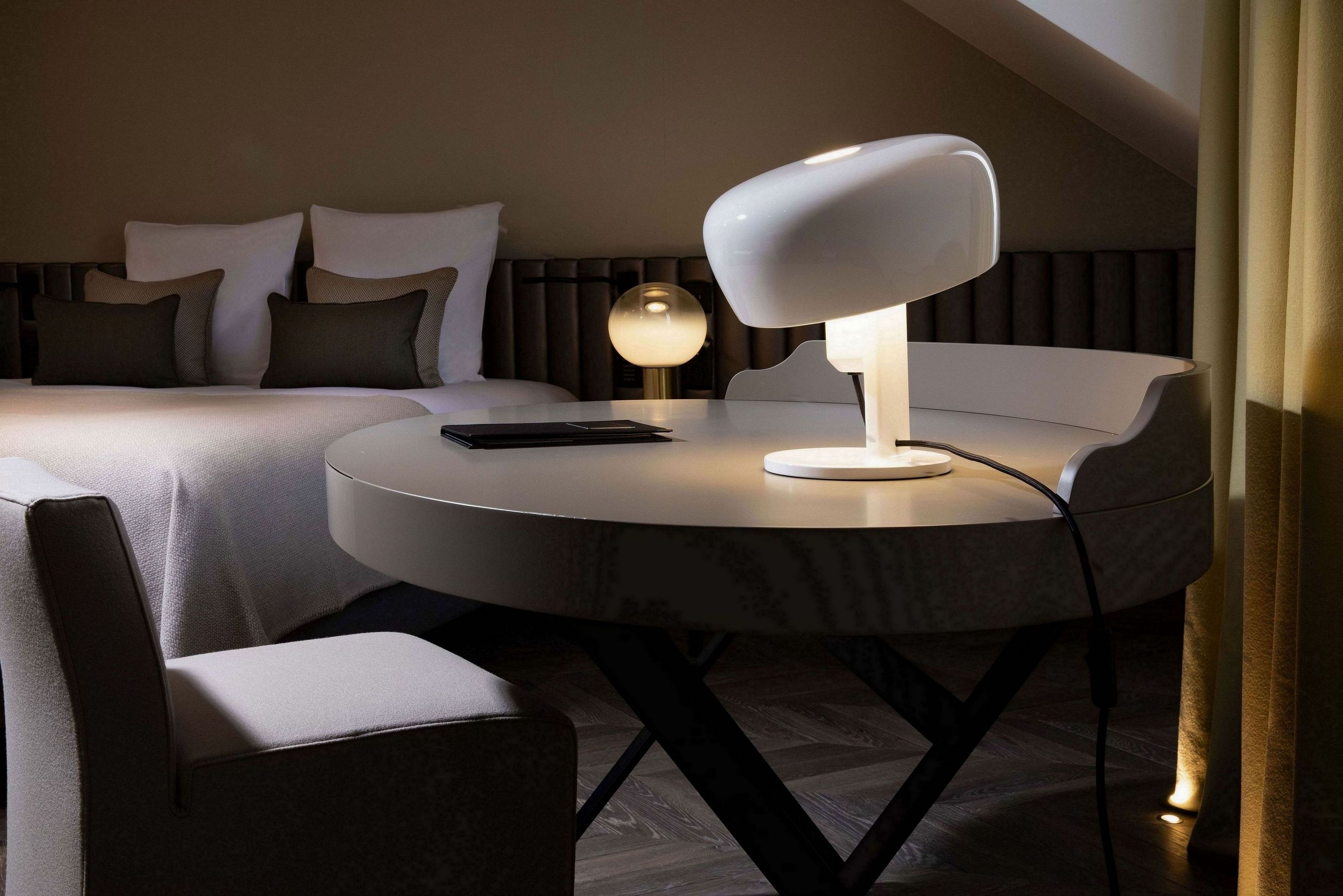 Une chambre d’hôtel moderne avec une table à manger ronde, une lampe blanche et un lit agrémenté de plusieurs coussins.