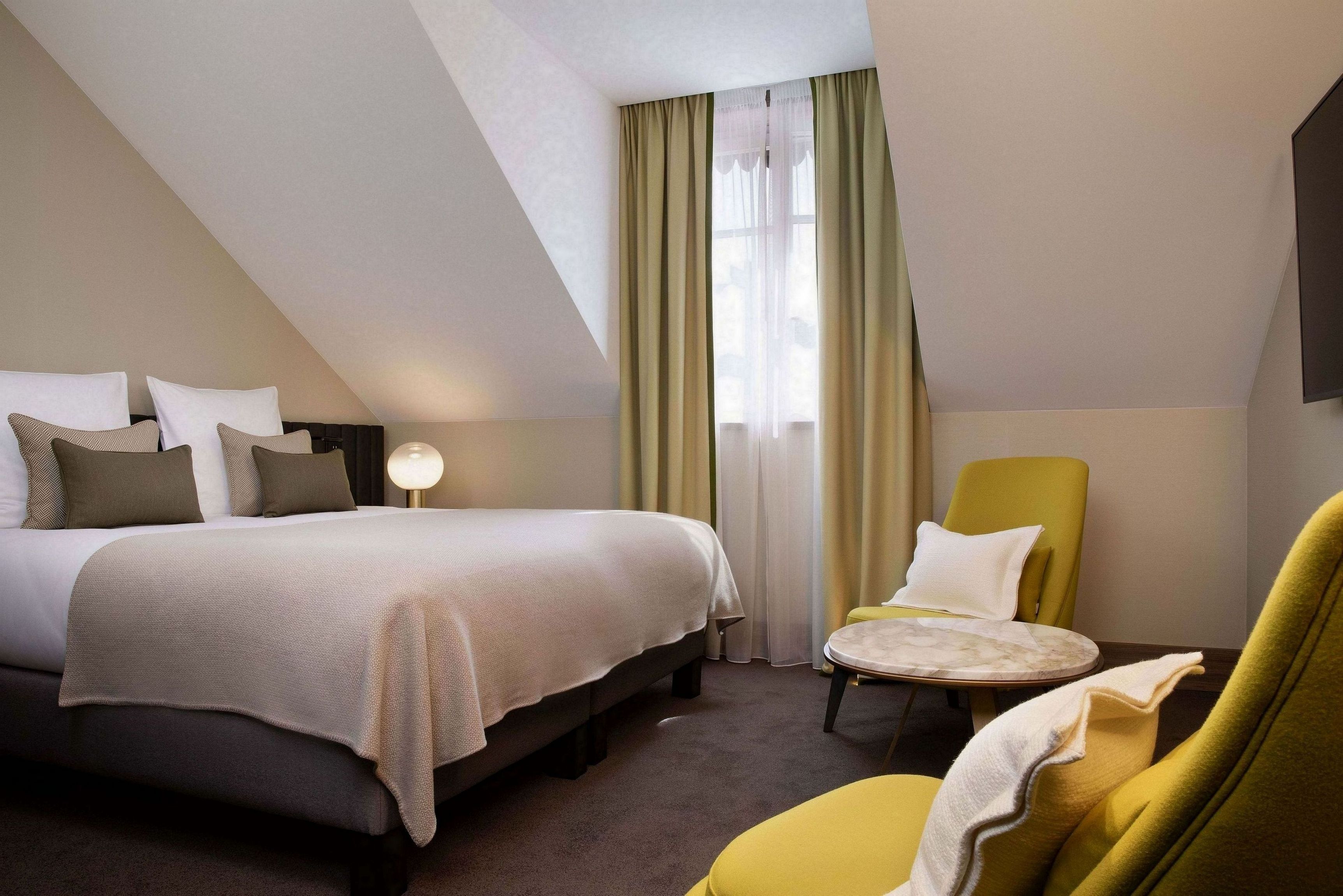 Ein Hotelzimmer mit einem Bett, einem runden Tisch, einem gelben Sessel und einem großen Fenster mit Vorhängen.