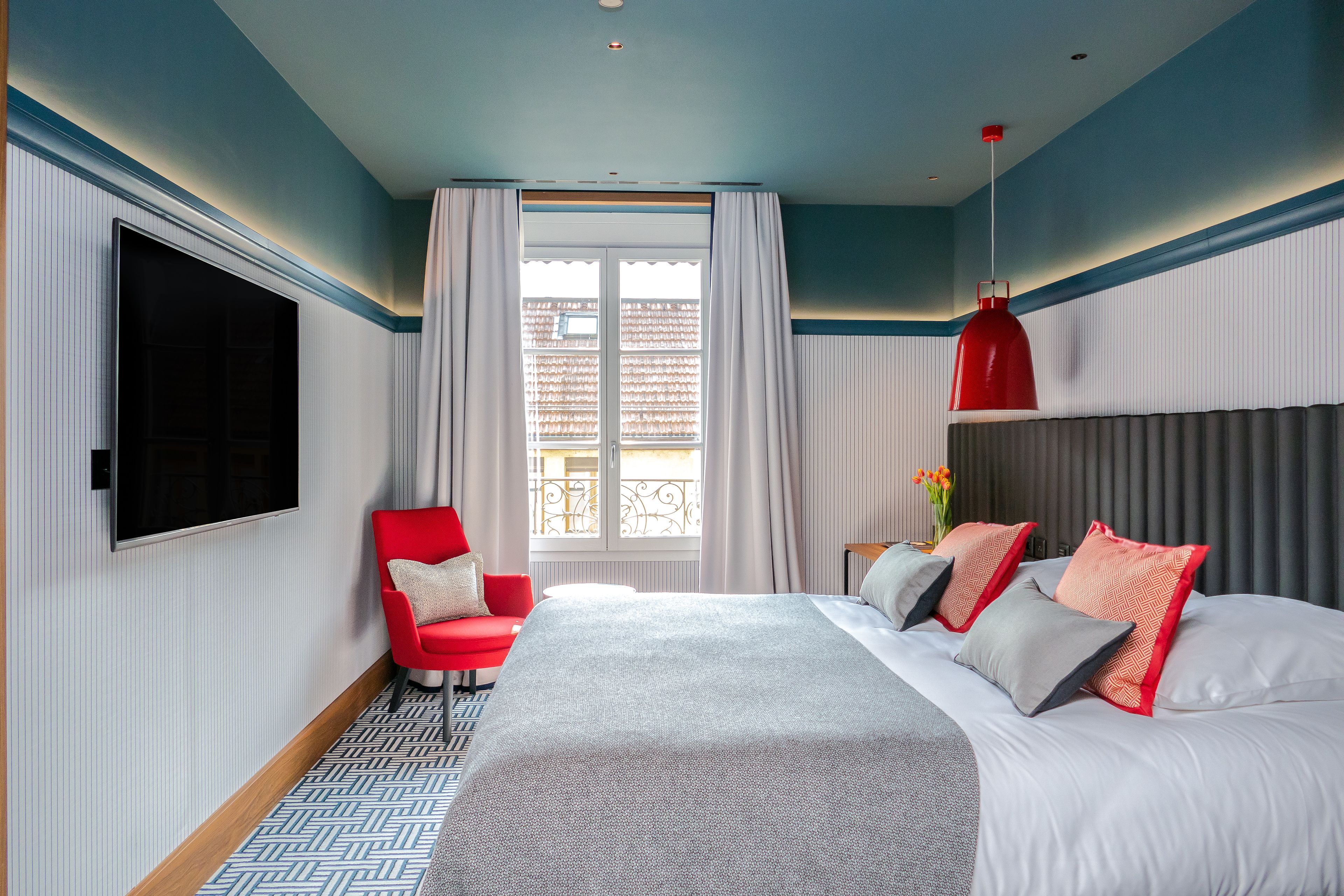A modern hotel room with a large bed, a red chair, a TV mounted on the wall, and a window with curtains.