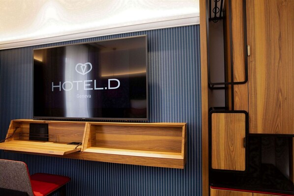 A modern hotel room with a large flat-screen TV mounted on a dark blue wall, wooden shelves below, and a red chair. - Hotel D Geneva (Geneva)