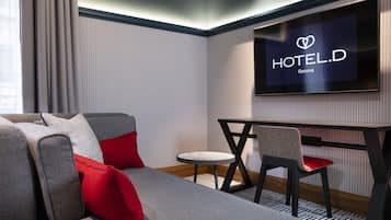 A modern hotel room with a sofa, red cushions, a small round table, and a TV displaying the HOTEL D Geneva logo.