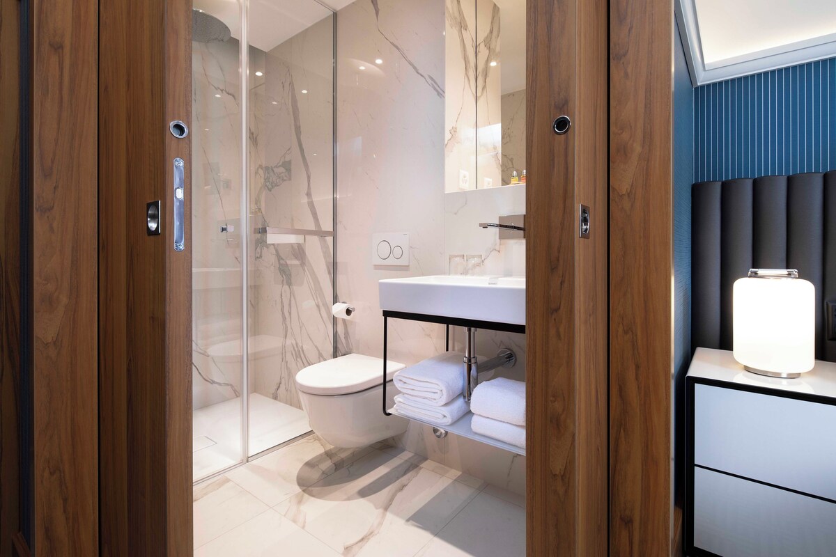 A modern bathroom with a glass shower enclosure, a white toilet, a vanity with a mirror, and a bedside table with a lamp.