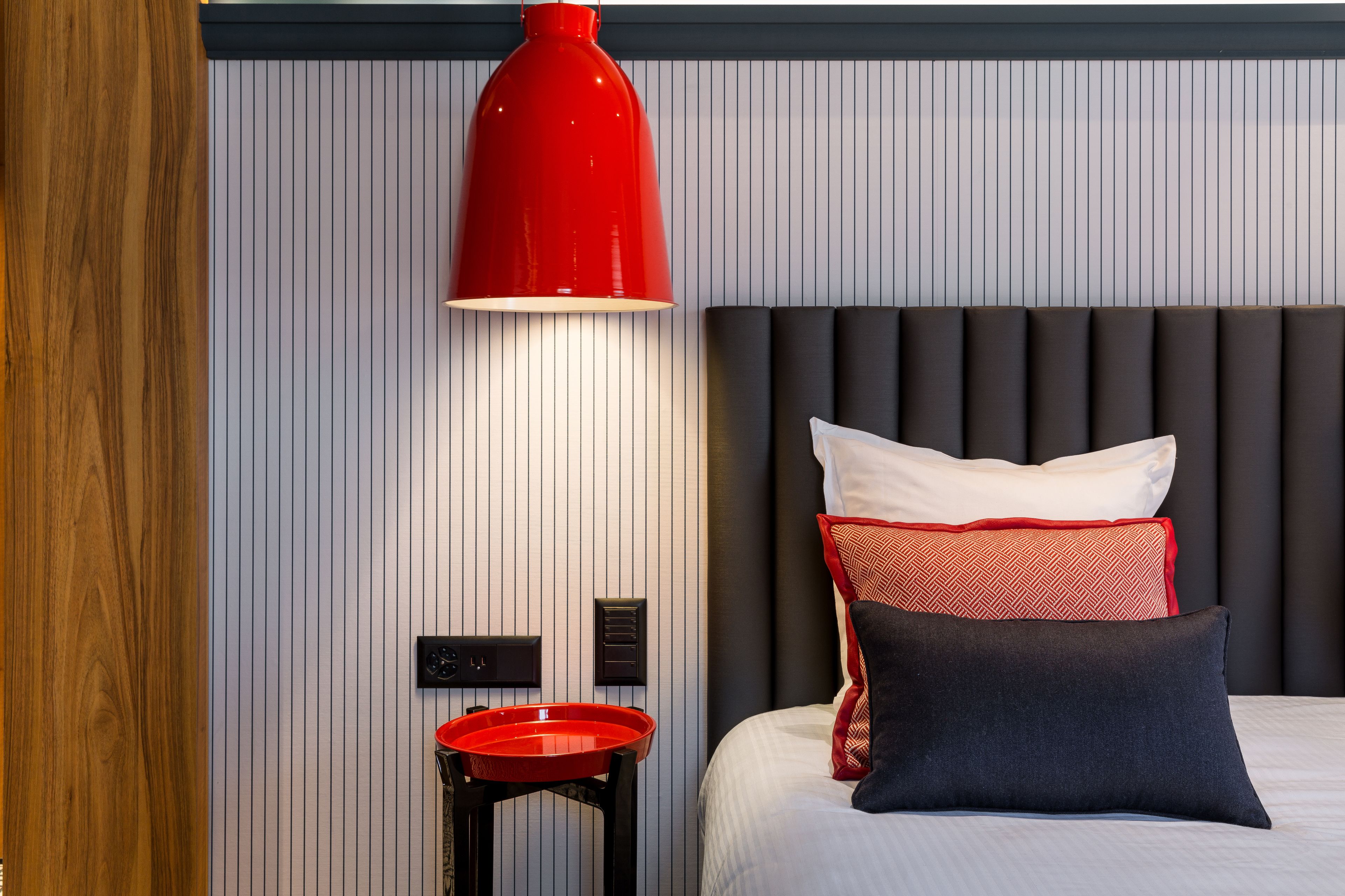 A modern hotel room with a large bed, a red pendant light, and a small red tray table.