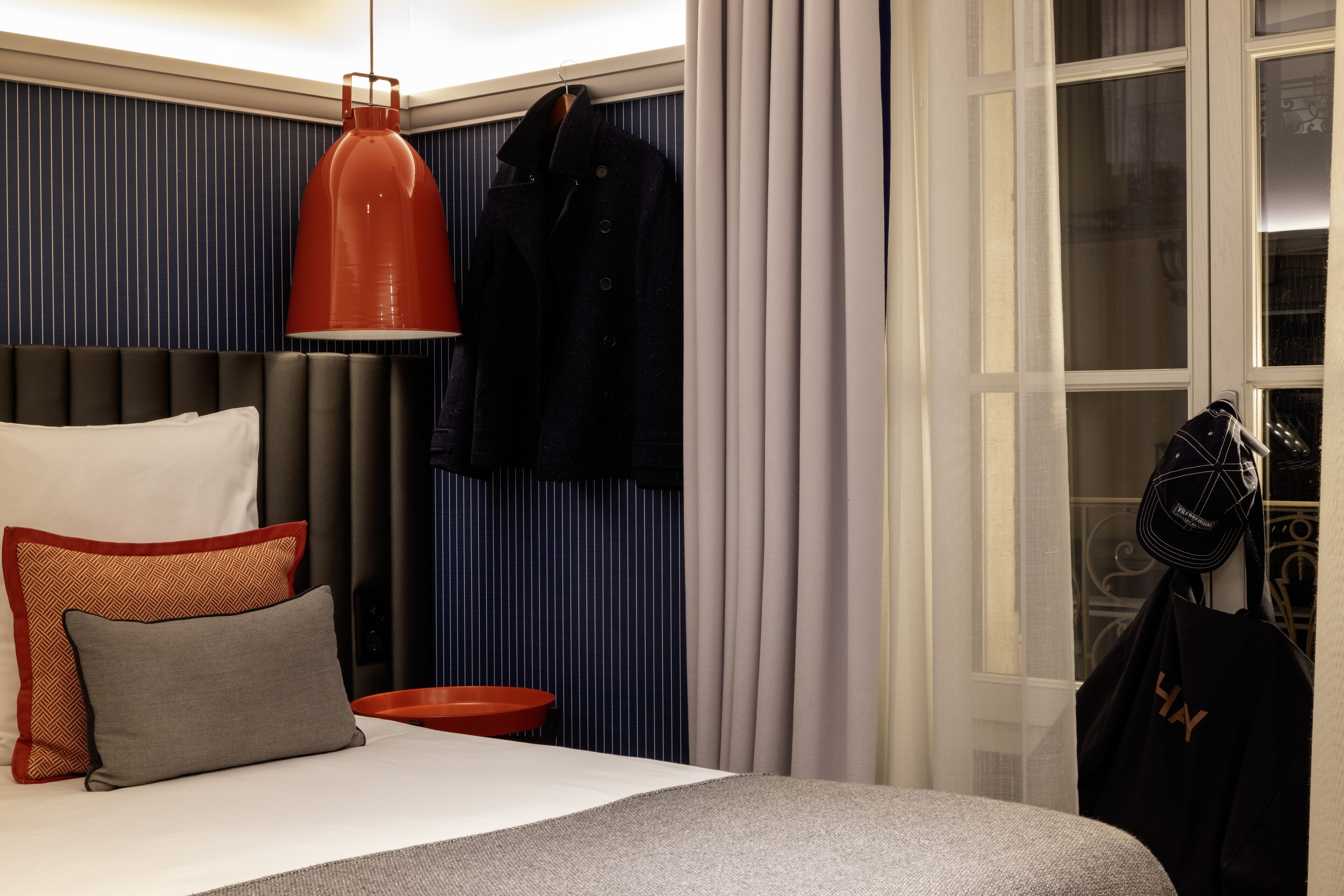 A hotel room with a bed, pillows, a red pendant light, a coat hanging, and a window with curtains.