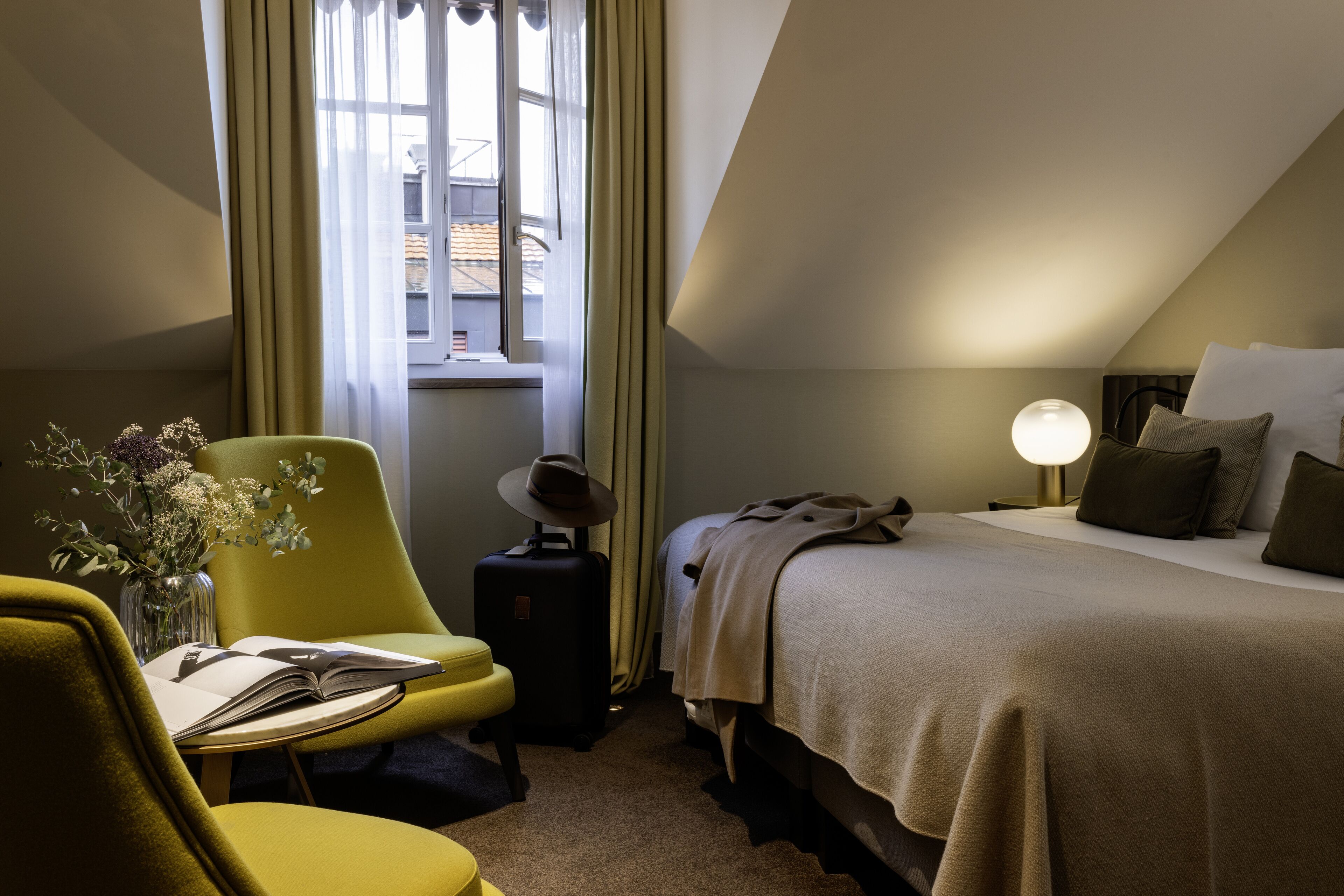 Ein Hotelzimmer mit Bett, Stuhl, Tisch mit einem Buch, Lampe und einem fensterbepflanzten Fenster.