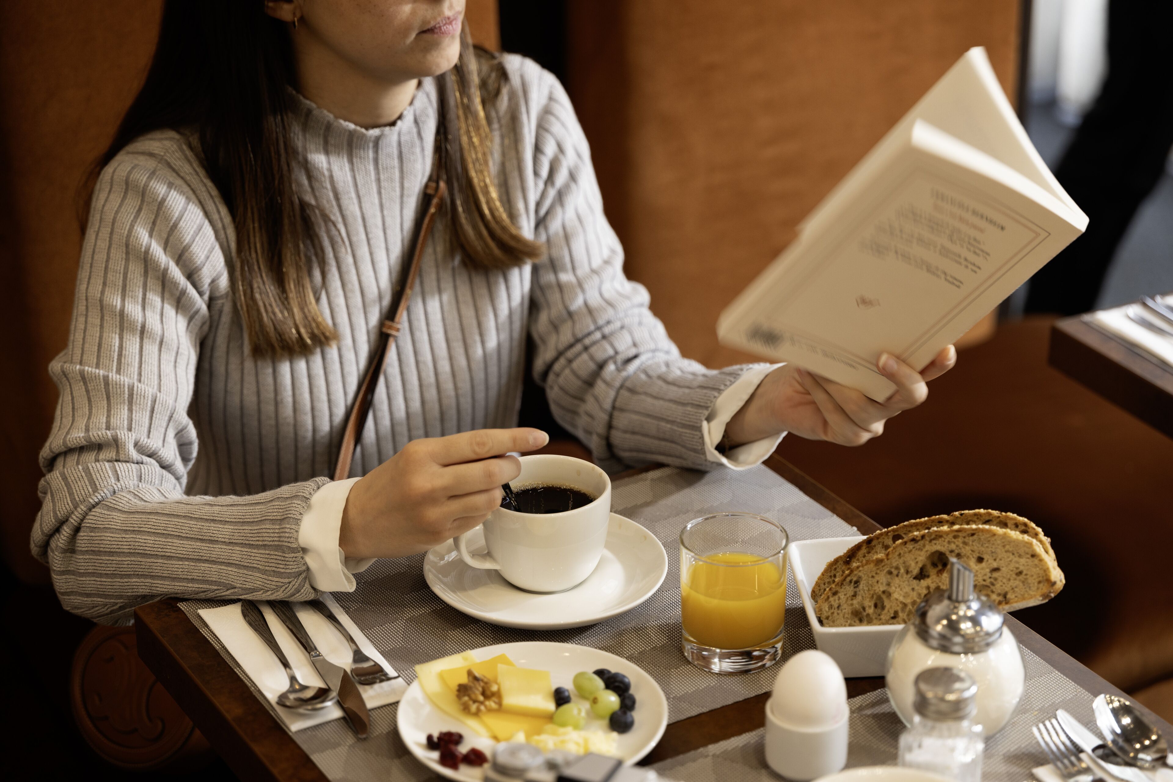 A person sitting at a table with a book, coffee cup, and breakfast plate including toast, fruit, and eggs.