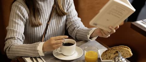A person sitting at a table with a book, coffee cup, and breakfast plate including toast, fruit, and eggs.