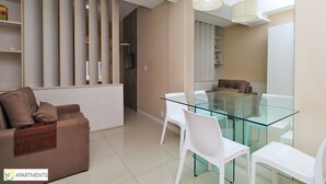 Ein modernes Wohnzimmer mit einem Glastisch, weißen Stühlen, einem braunen Sofa und einer beige gefärbten Wand mit vertikalen Jalousien.