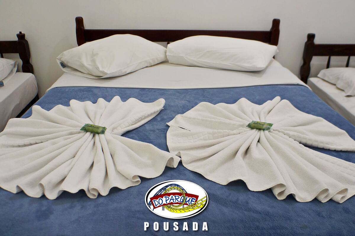 A hotel room with two beds, each with white pillows and blue bedspreads. There are two towels folded on the beds, and a logo with the text "DO PARQUE" and "POUSADA" is visible on the bedspread.