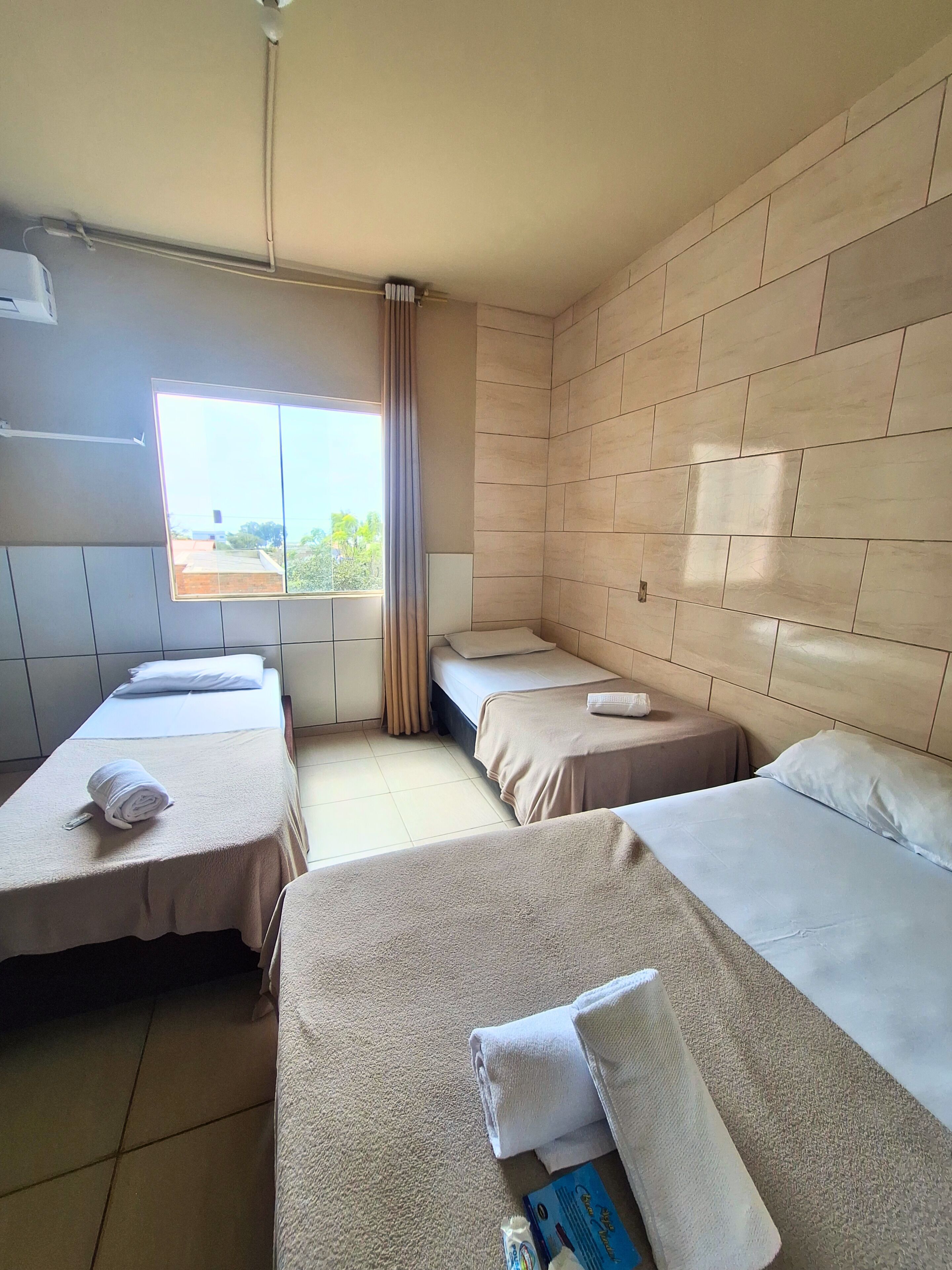 A hotel room with three single beds, a window with curtains, and a view of the outside.