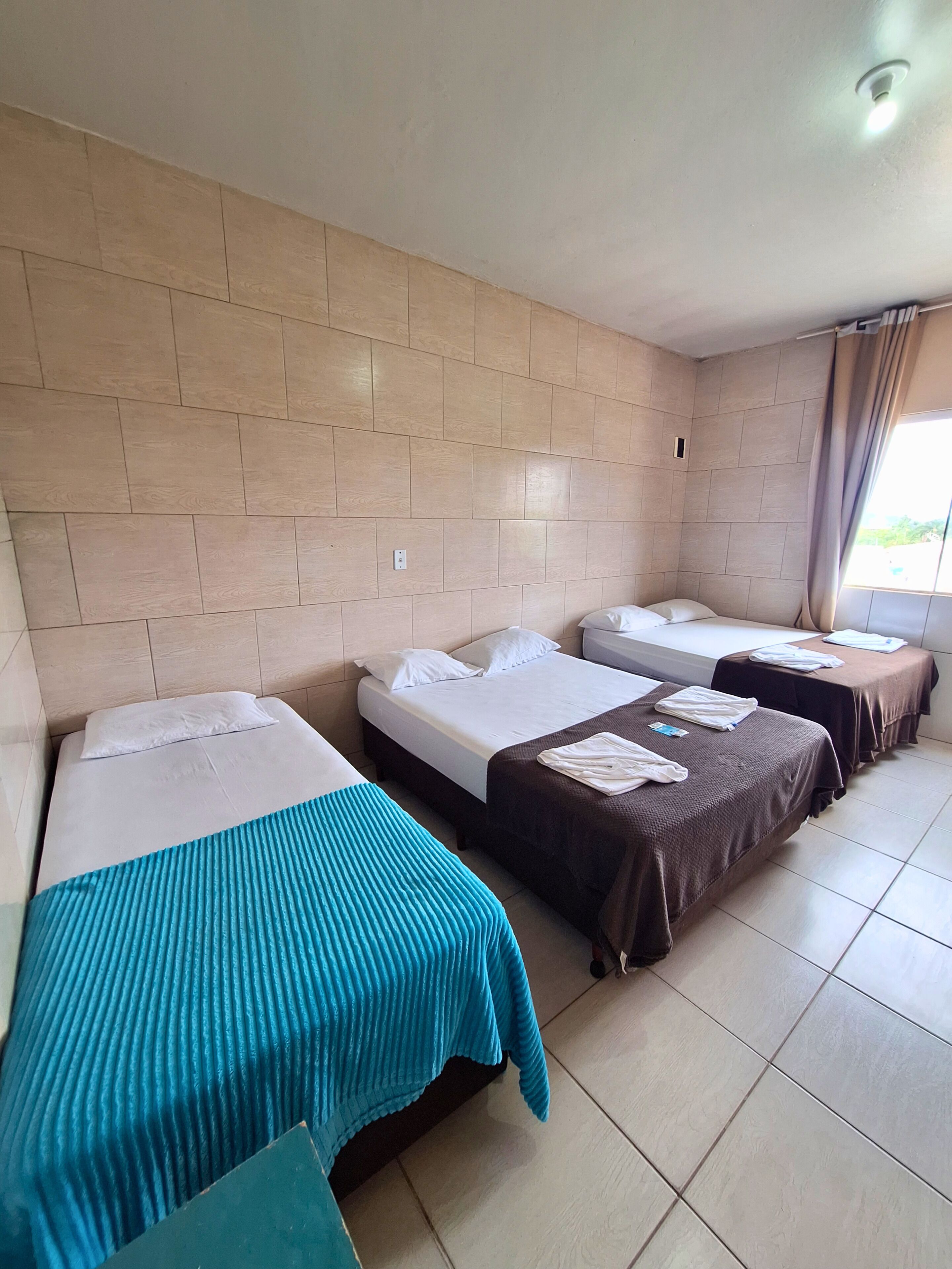 Una camera d'albergo con tre letti allineati, una finestra con tende e un pavimento piastrellato.