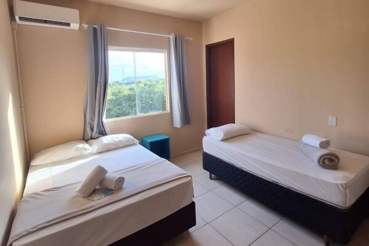 A room with two beds, a window with curtains, an air conditioning unit, and a door.