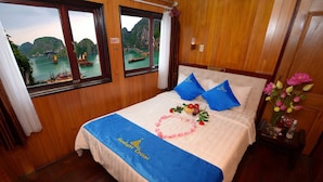 Habitación de hotel con cama, paredes de madera, dos ventanas con vistas a barcos y montañas, y una mesita de noche con un jarrón de flores.
