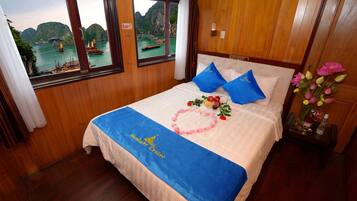 A hotel room with a bed, wooden walls, two windows with a view of boats and mountains, and a nightstand with a vase of flowers.