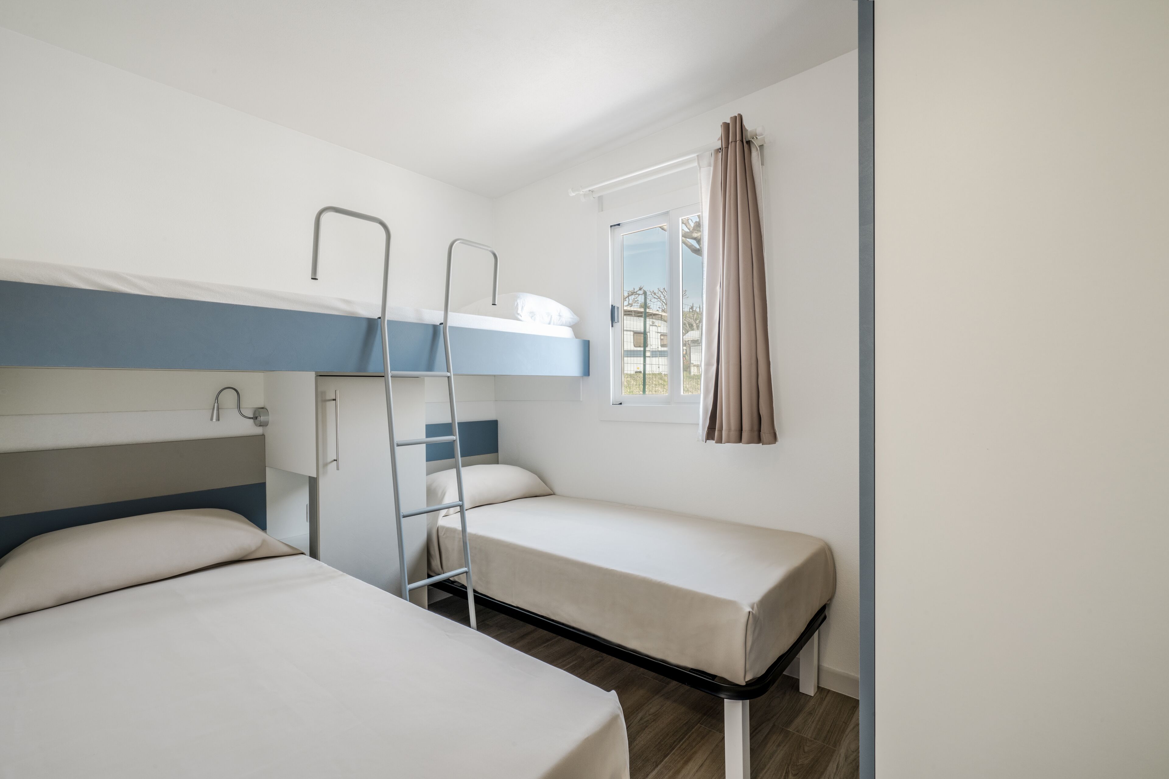 A dormitory room with bunk beds, a ladder, a window with curtains, and a view of the outside.