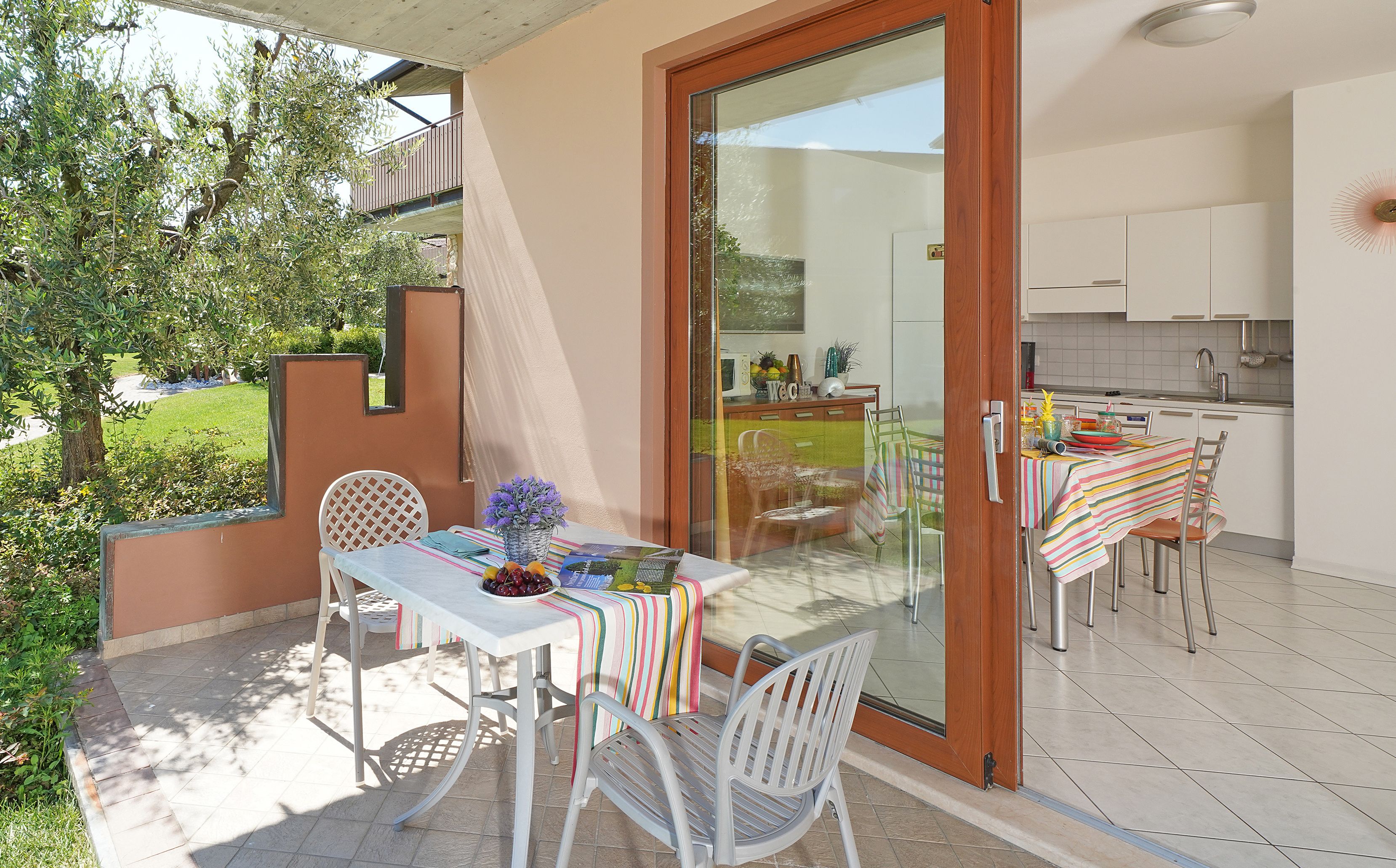 An outdoor dining area with a table set for two, a sliding glass door leading to a kitchen, and a view of a garden.