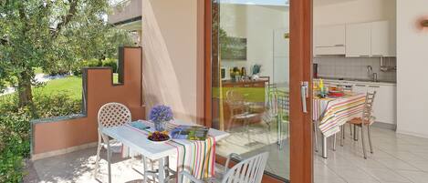 An outdoor dining area with a table set for two, a sliding glass door leading to a kitchen, and a view of a garden.
