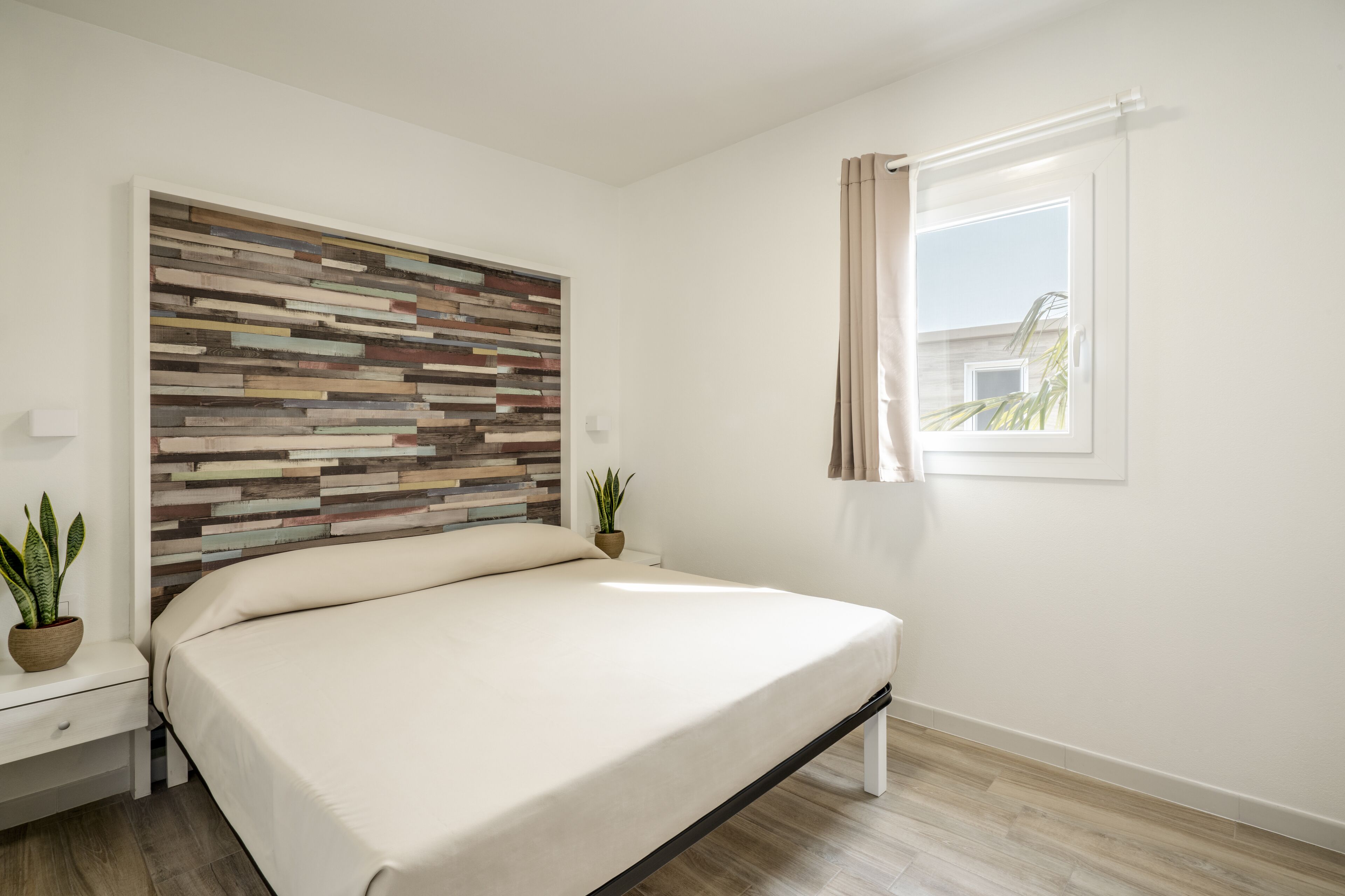A bedroom with a bed, a nightstand, a potted plant, and a window with curtains.