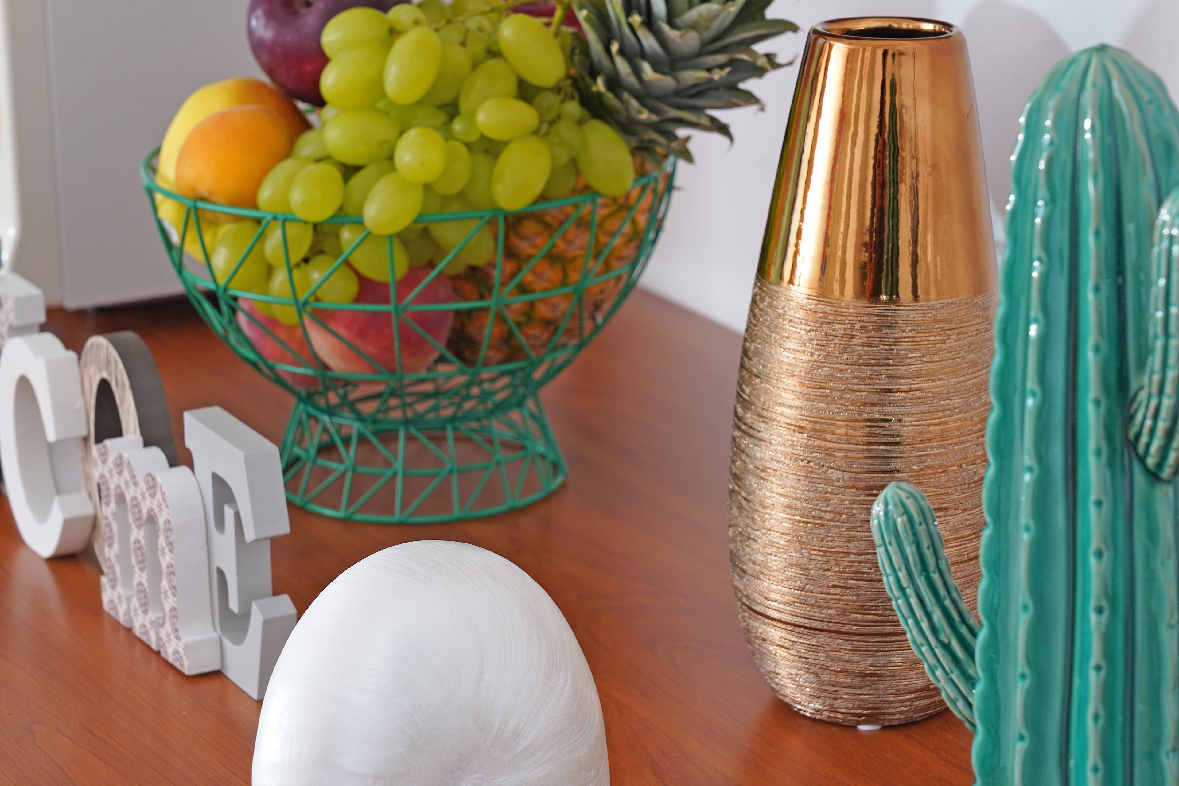 A fruit bowl with grapes and pineapple, a golden vase, and a green cactus on a wooden table.