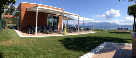 A modern outdoor seating area with tables and chairs, a wooden building, and a view of a lake and mountains.