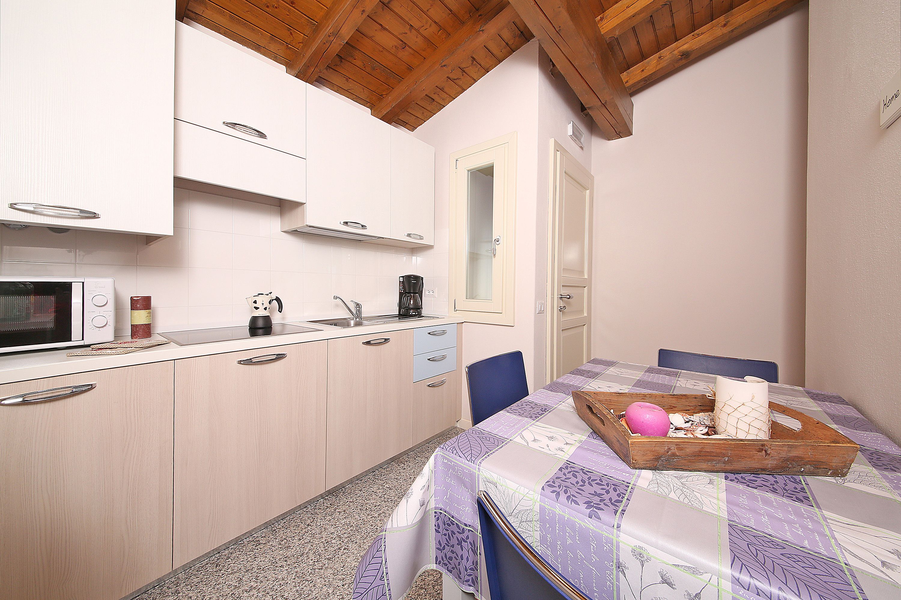 A compact kitchen with white cabinets, a microwave, and a coffee maker. There's a dining table with a purple tablecloth and a wooden tray with a pink ball and a candle.