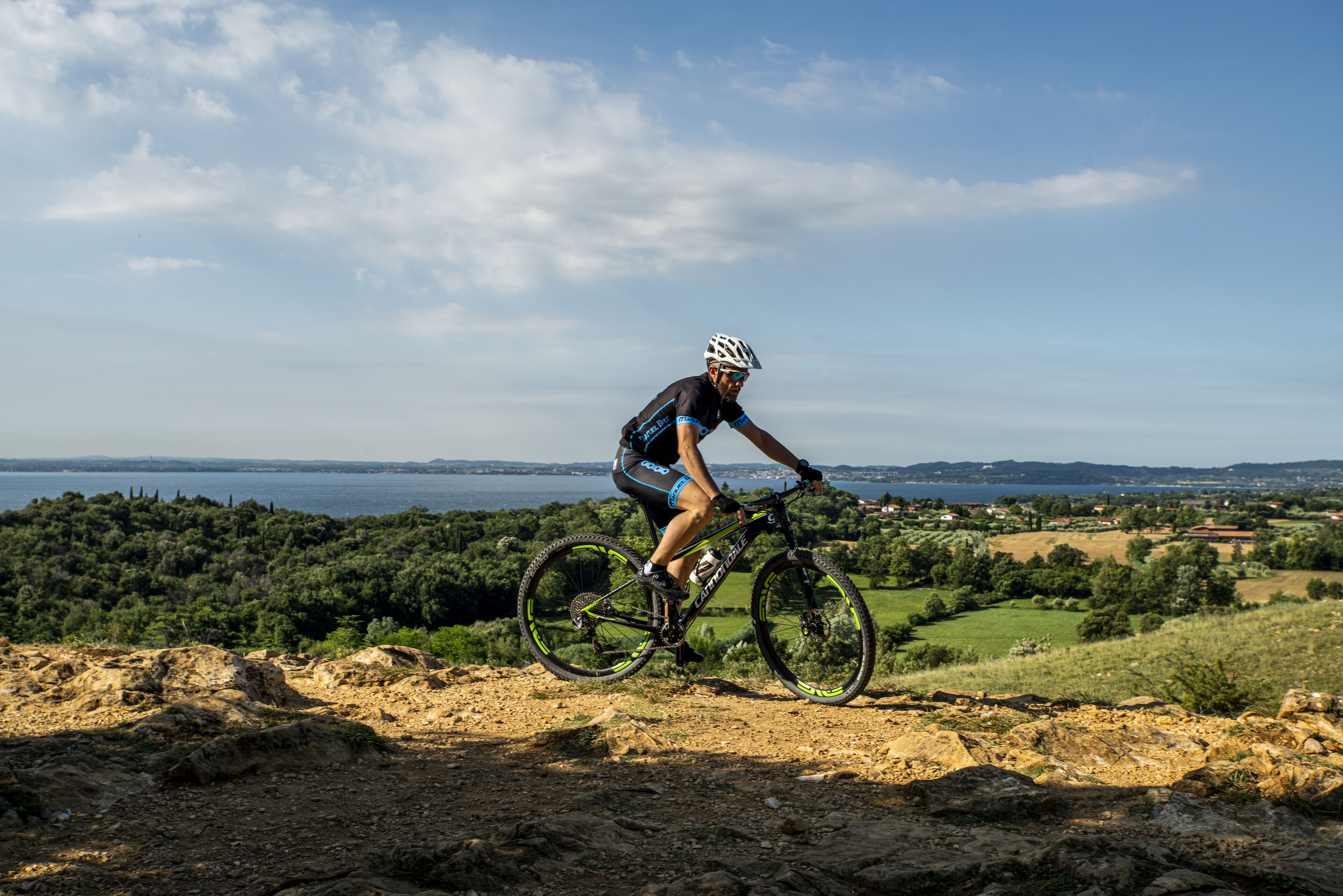 A cyclist on a mountain bike trail with a scenic view of a lake and hills in the background.