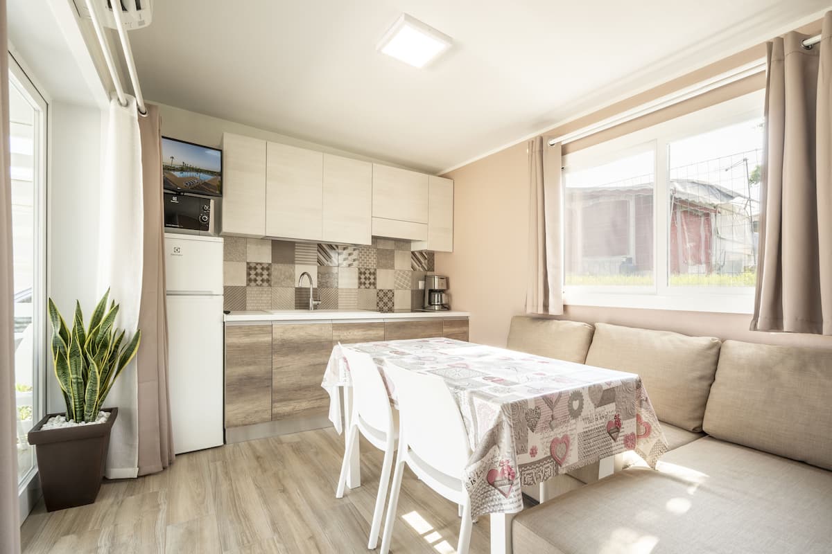 Eine moderne Küche mit weißen Schränken, integriertem Mikrowellenherd und einer Kaffeemaschine. Ein Esstisch mit gemustertem Tischläufer und weiße Stühle bieten Platz. Ein großes Fenster sorgt für natürliches Licht und einen Ausblick nach draußen.