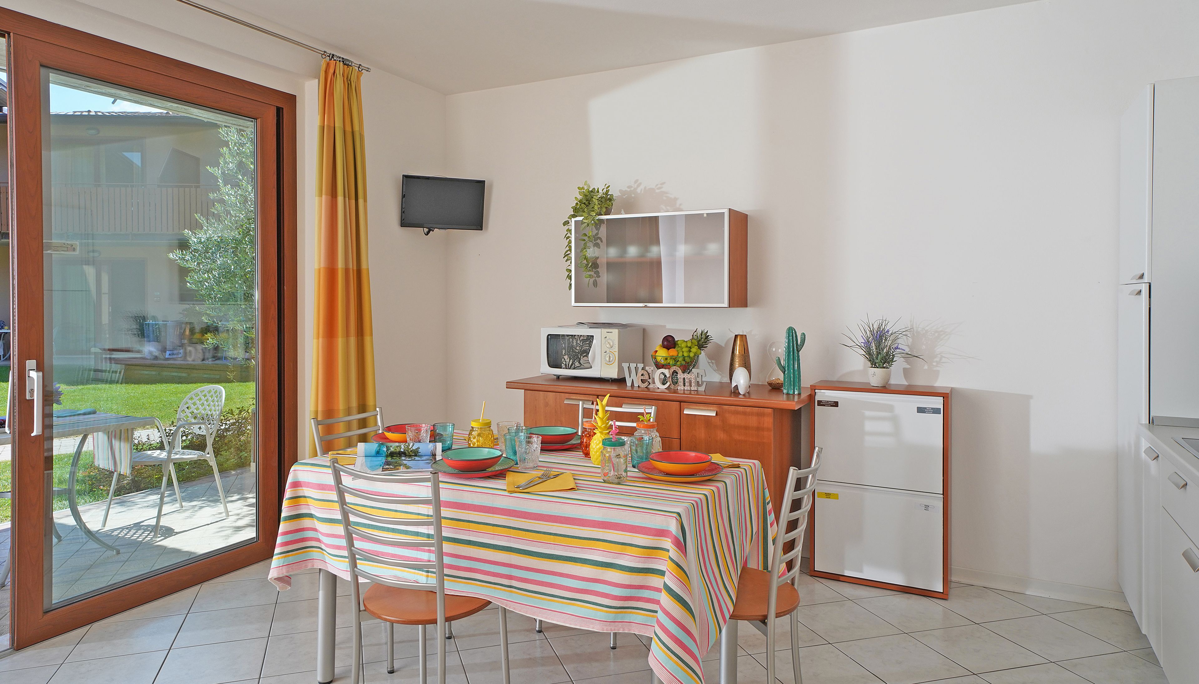 Une petite cuisine équipée d’un réfrigérateur, d’un four à micro-ondes et d’un petit placard. On y trouve une table à manger avec des chaises et une nappe à rayures. Un téléviseur est fixé au mur, au-dessus du plan de travail de la cuisine.