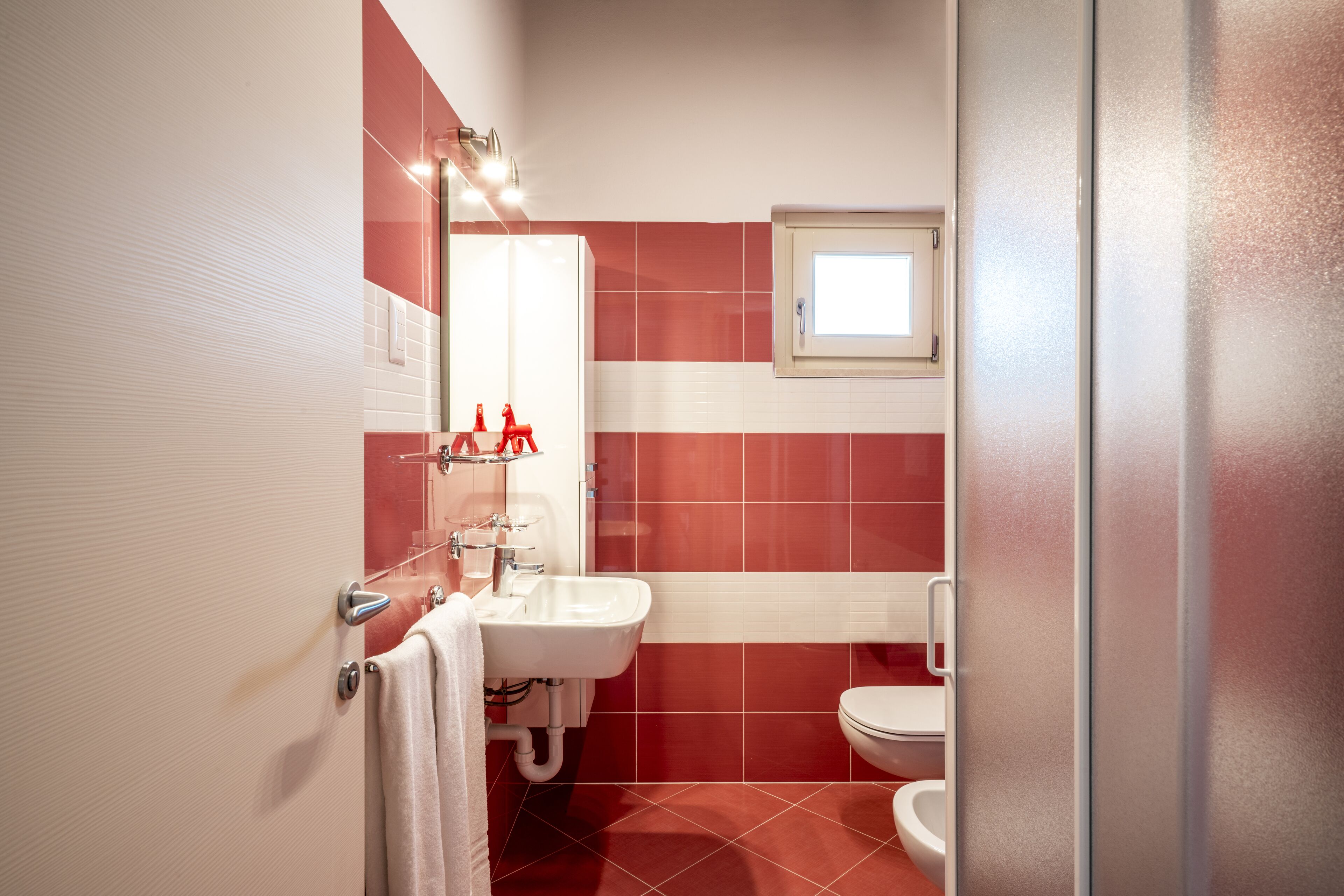 Un baño con un lavamanos blanco, azulejos rojos, un inodoro blanco y una ventana sobre el inodoro.