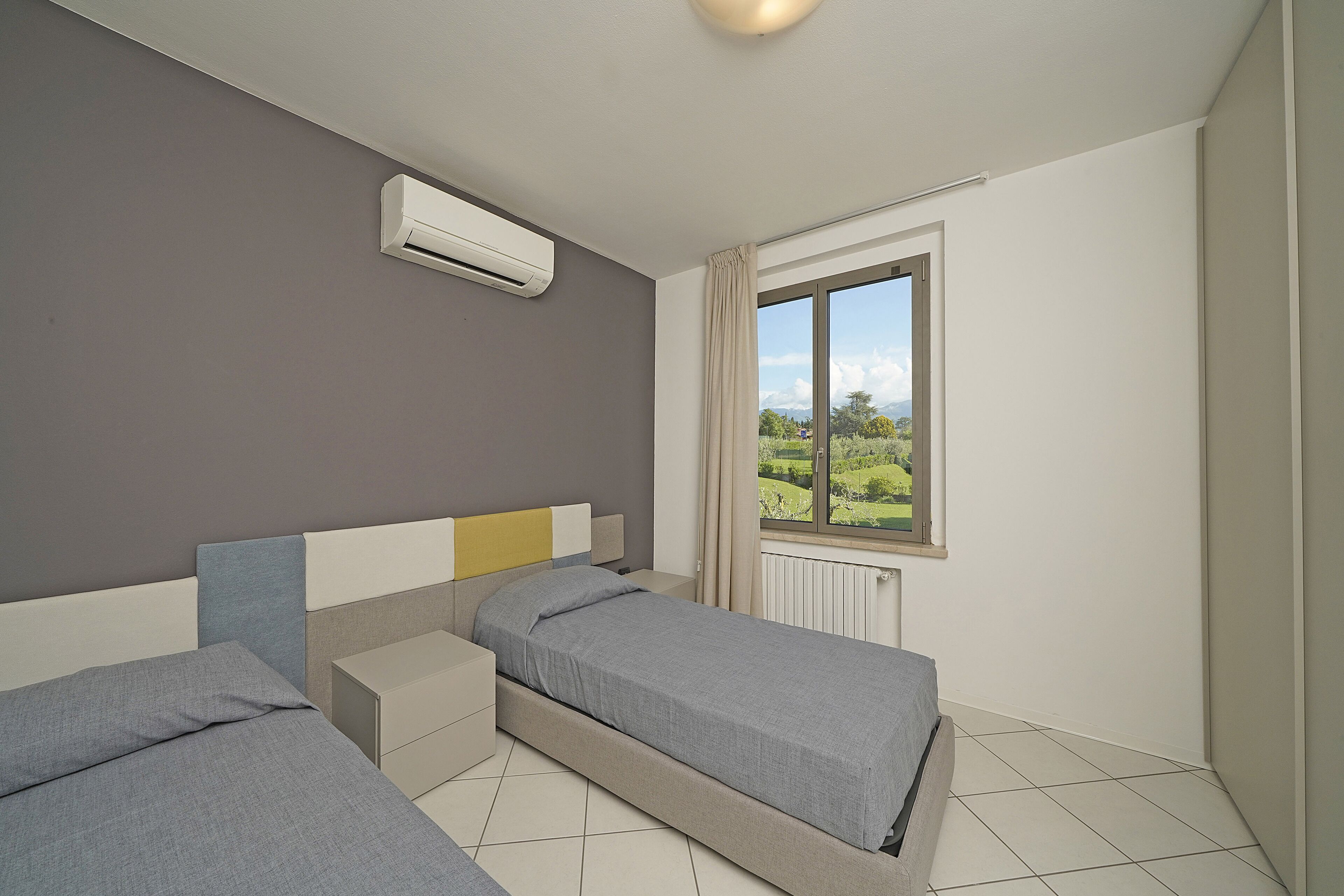 Un dormitorio con dos camas individuales, una ventana con vistas a zonas verdes y un equipo de aire acondicionado en la pared.