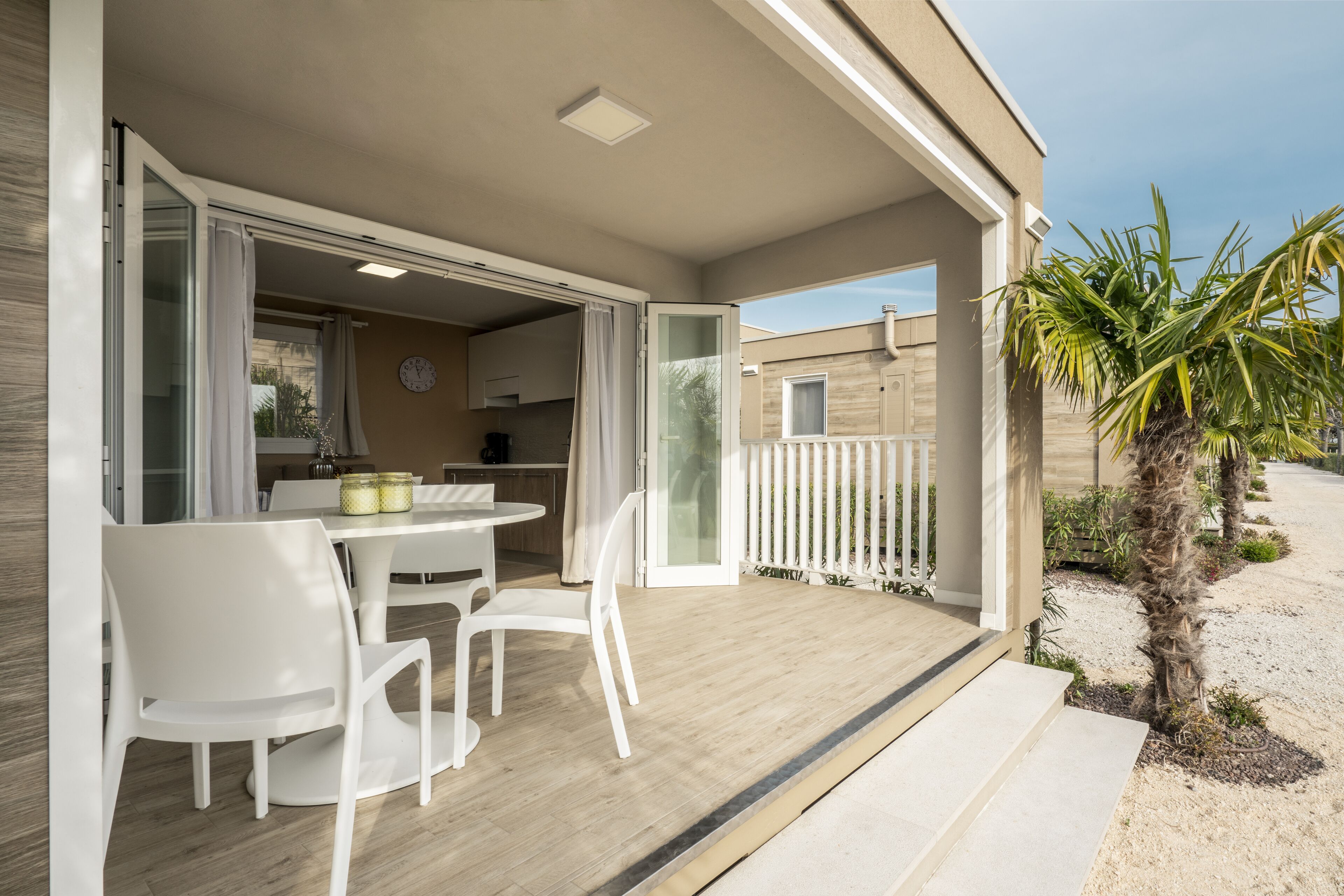 Une terrasse ouverte avec une table et des chaises, une porte-fenêtre coulissante donnant sur une cuisine et un petit jardin agrémenté d’un palmier.