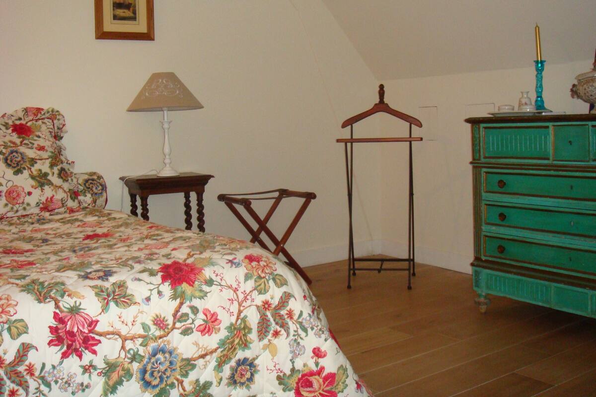 Un dormitorio con colcha estampada, una silla de madera, un aparador y una percha.