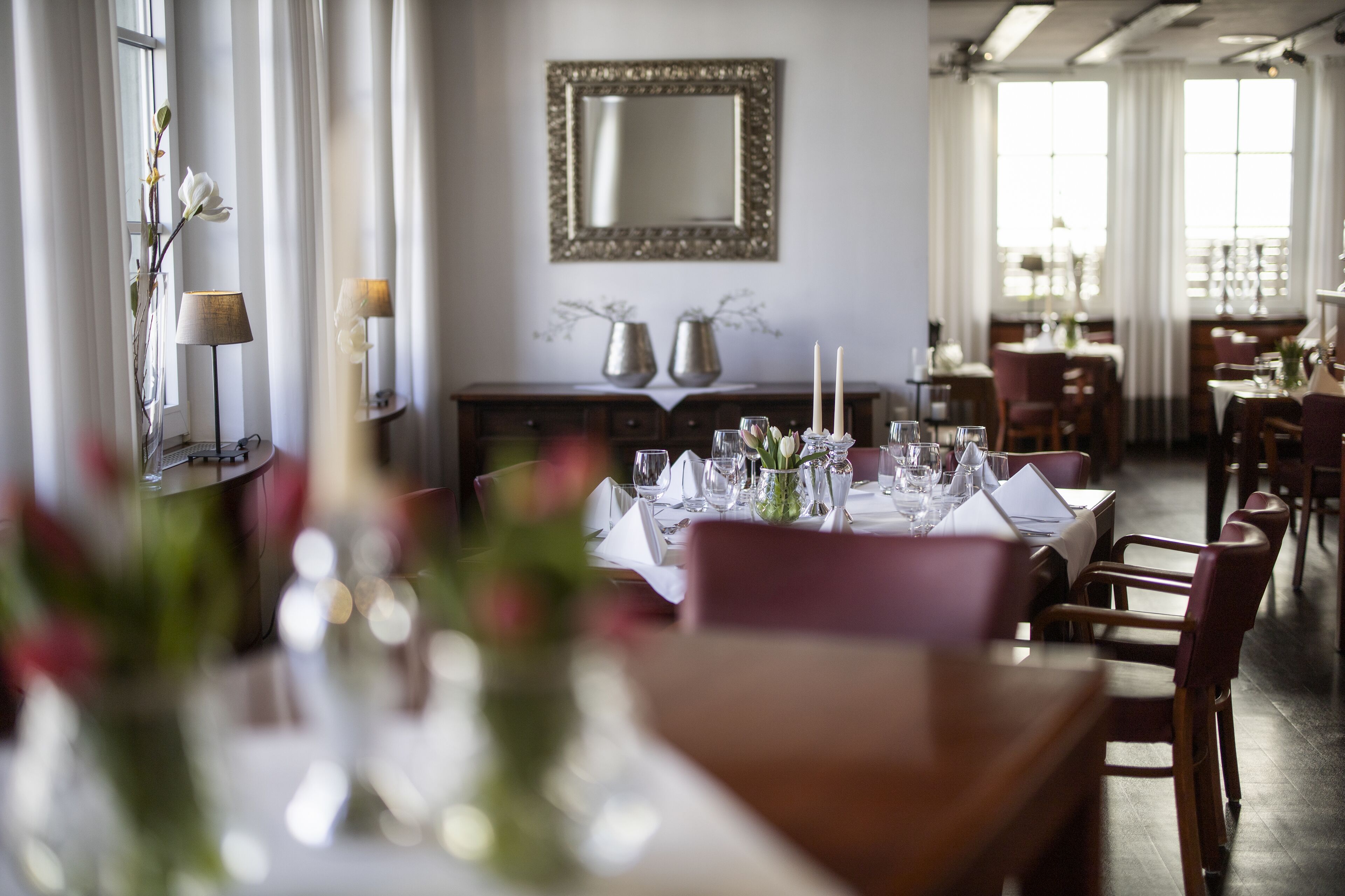 A dining area with tables set for a meal, a mirror on the wall, and a window with curtains.