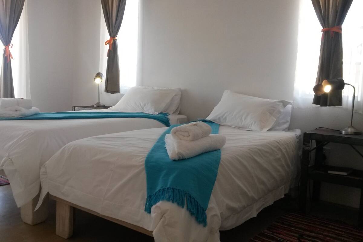 A hotel room with two beds, a nightstand, a lamp, and a window with curtains.