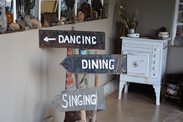 A rustic interior with a vintage stove, wooden signposts labeled 'DANCING,' 'DINING,' and 'SINGING,' and a shelf with various bottles and items.