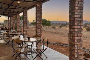 Un área de comedor al aire libre con pilares de ladrillo, mesas y sillas de madera, con vistas a un paisaje desértico.