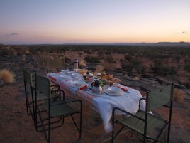 Mesa para comer al aire libre con sillas, platos y una botella, rodeada por un paisaje desértico.
