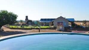 A swimming pool with a view of a building with solar panels, a small shed, and a grassy area with a tree. - Alte Kalkofen Lodge (Simplon)