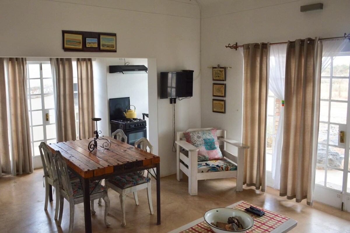 A dining area with a wooden table and chairs, a television mounted on the wall, and a window with curtains.