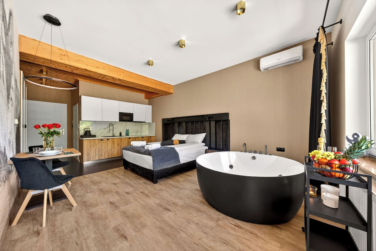 A modern hotel room with a large bathtub, a dining area, and a kitchen.
