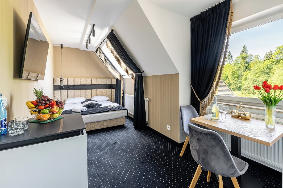 A modern hotel room with a bed, a dining table, a chair, a fruit bowl, and a view of trees.
