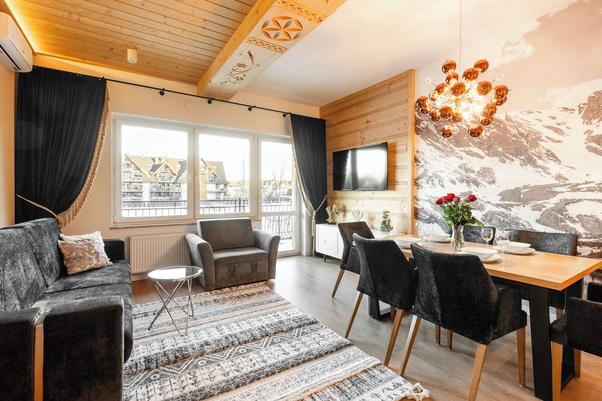 A modern living room with a sofa, chairs, a dining table, and a TV mounted on the wall.