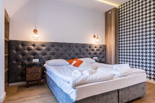 Una cama bien hecha con ropa de cama blanca y un cabecero gris, una mesita de noche de madera con cajones, y un papel tapiz estampado en la pared derecha.
