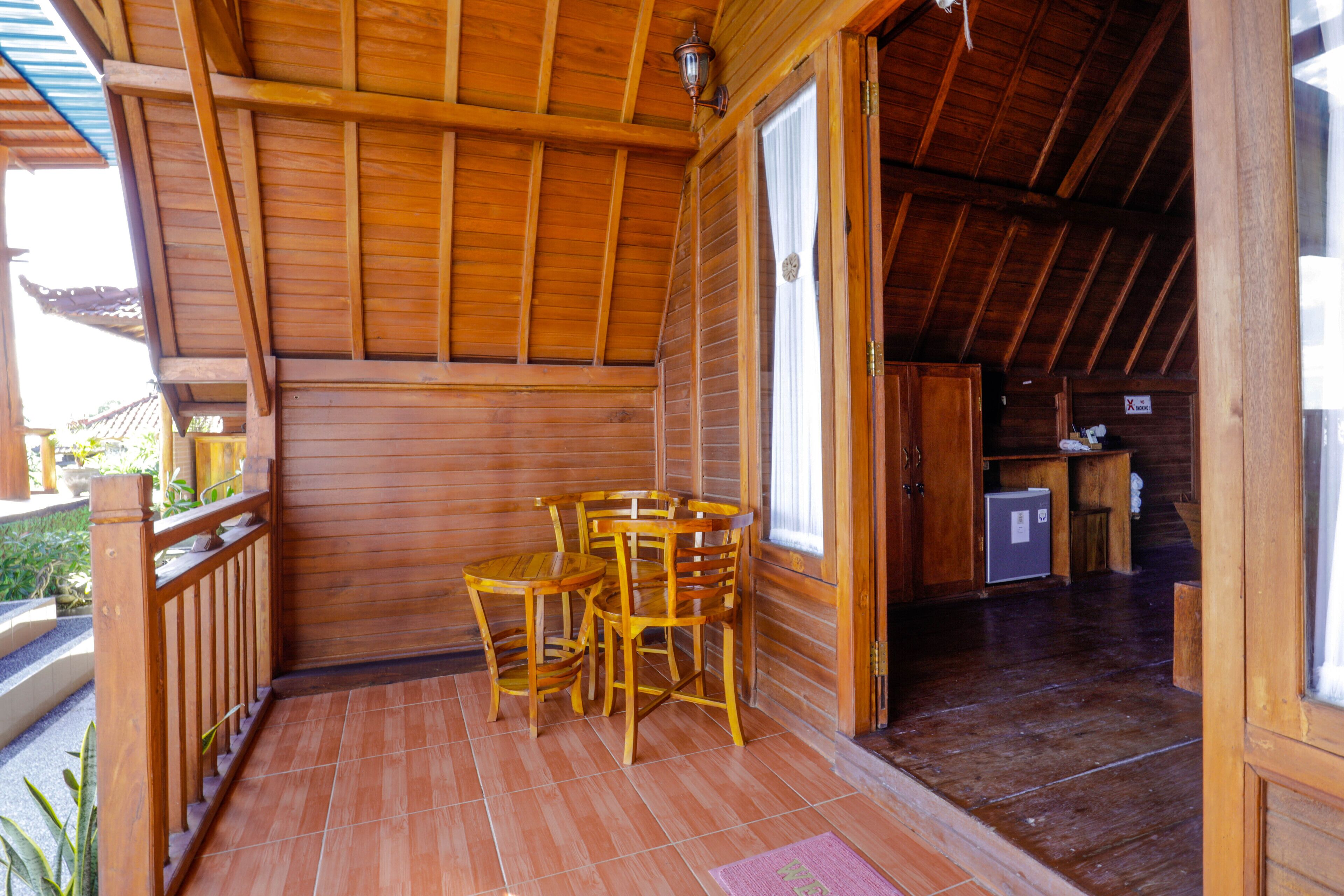 A wooden cabin with a porch, a dining area with chairs and a table, and a kitchen area with a refrigerator.