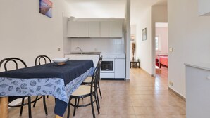 Cocina compacta con armarios blancos, una mesa de comedor con sillas y vistas a un dormitorio con colcha roja.