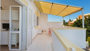 Un espacio al aire libre cubierto con un toldo blanco y amarillo a rayas, una puerta blanca que da a una habitación con escritorio y silla, y vistas de otros edificios al fondo.